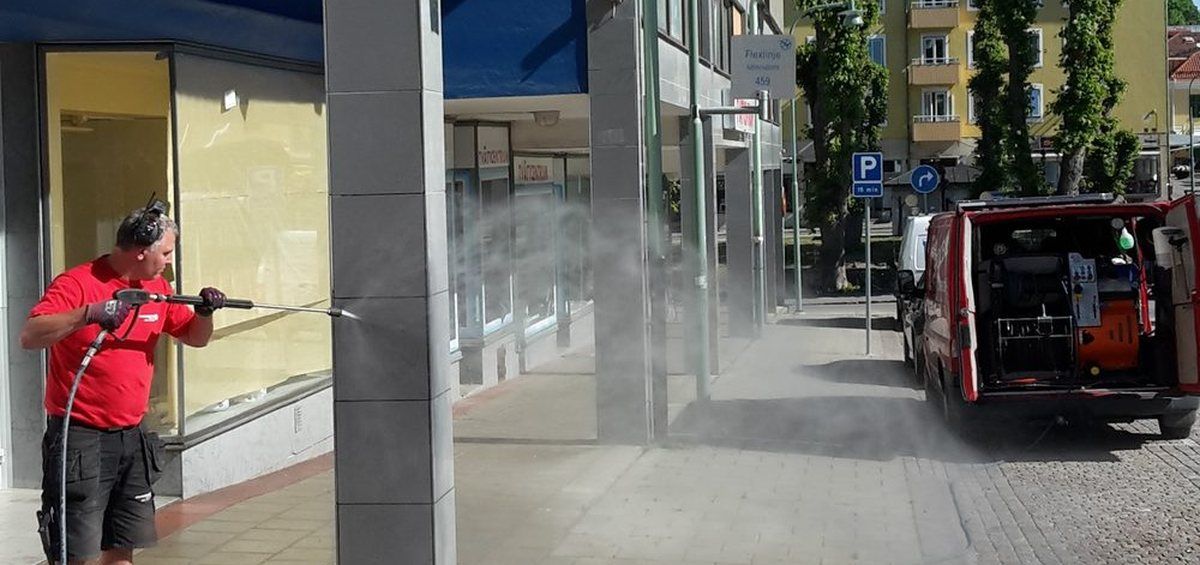 Medarbetare i röd tröja hetvattenspolar en gata utanför en butikslokal, med den röda servicebilen och utrustningen bakom sig.